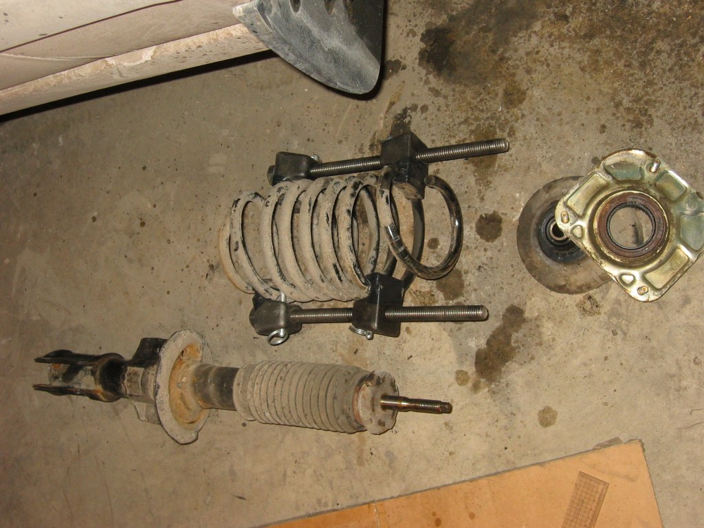 Do NOT unscrew the top retaining nut or the cross shaped retaining nut.  Or else, the spring will fly right out and cause some serious damage.  Mount the spring compressor according to its instructions and compress the spring.  For spring compressors like the ones shown, put them 180 degrees across and turn both sides evenly.  Compress until the spring can wiggle between the spring seat and the bottom of the strut.